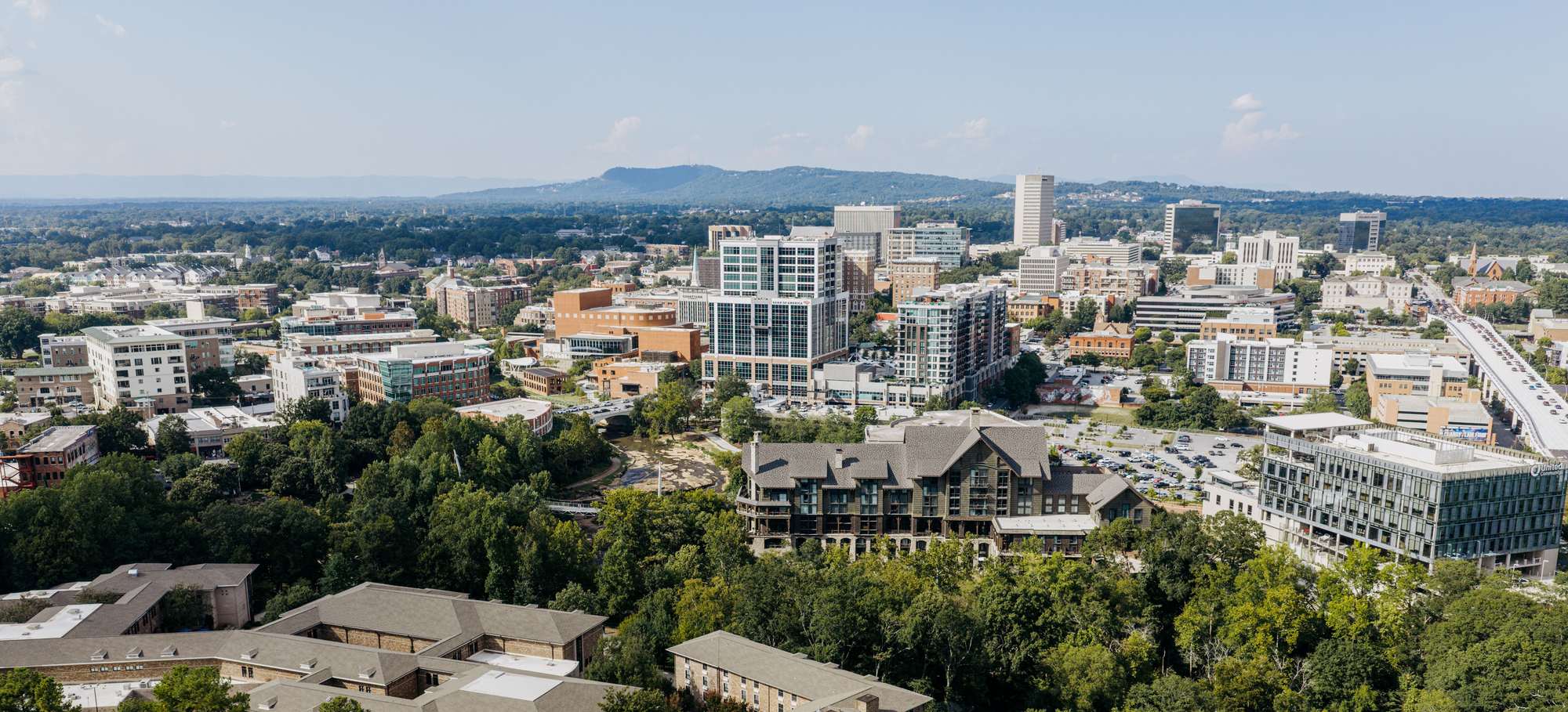 Downtown Greenville seen from the south, with the wooded Reedy River corridor in the foreground and the Blue Ridge on the horizon.