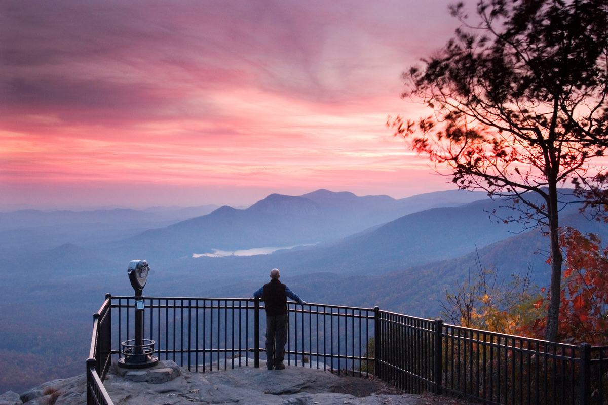 A visitor at the overlook railing at Caesars Head, looking out over the Blue Ridge under a pink sky.